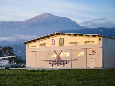A large, light-colored aircraft hangar featuring logos for aviation brands like Garmin, Cessna, and Diamond, with a large mural of a propeller plane on the door and the text "AEROSERVICE" and "WWW.AEROSERVICE.SI" visible on the facade.