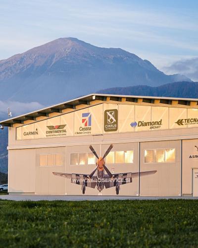A large, light-colored aircraft hangar featuring logos for aviation brands like Garmin, Cessna, and Diamond, with a large mural of a propeller plane on the door and the text "AEROSERVICE" and "WWW.AEROSERVICE.SI" visible on the facade.