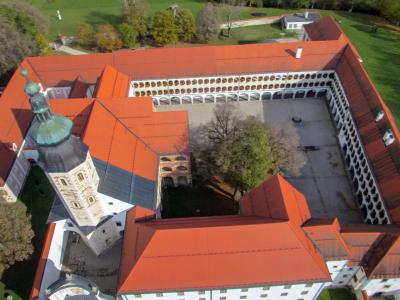 An aerial view of a large monastery complex with red-tiled roofs, a prominent church tower with a green copper spire, and a central courtyard surrounded by arcades.