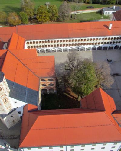 An aerial view of a large monastery complex with red-tiled roofs, a prominent church tower with a green copper spire, and a central courtyard surrounded by arcades.