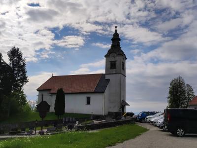 A white church with a red-tiled roof and a tall, dark-shingled tower stands on a grassy hill, surrounded by a cemetery and a parking lot with several cars under a cloudy blue sky.