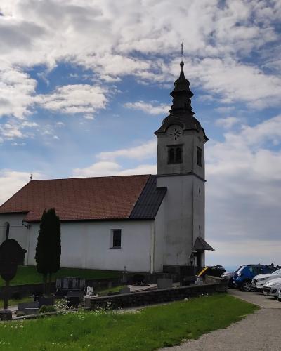 A white church with a red-tiled roof and a tall, dark-shingled tower stands on a grassy hill, surrounded by a cemetery and a parking lot with several cars under a cloudy blue sky.