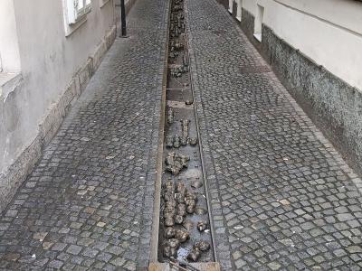 A long, narrow cobblestone alleyway featuring a central stone trough filled with numerous small, clustered bronze sculptures of faces and heads.