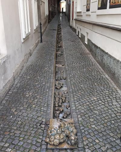 A long, narrow cobblestone alleyway featuring a central stone trough filled with numerous small, clustered bronze sculptures of faces and heads.