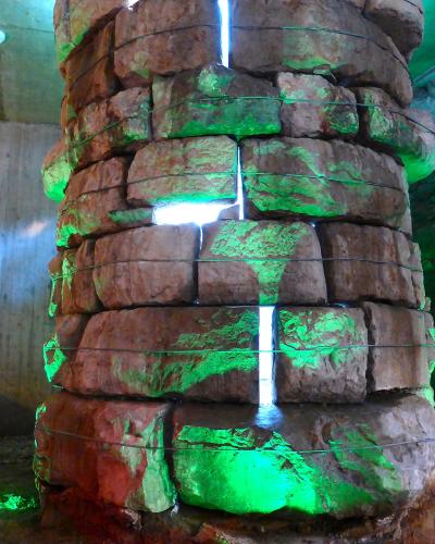 A cylindrical stone structure, likely a historic well or foundation pillar, illuminated with green and white lights inside a subterranean or basement-like space.