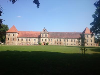 A long, two-story historical building with a central gable and two corner towers with red-tiled roofs, set behind a large green lawn under a blue sky.