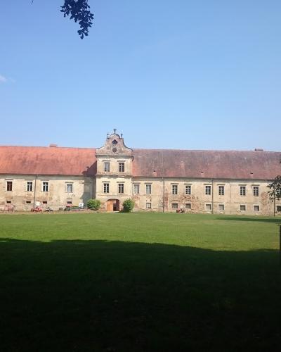 A long, two-story historical building with a central gable and two corner towers with red-tiled roofs, set behind a large green lawn under a blue sky.