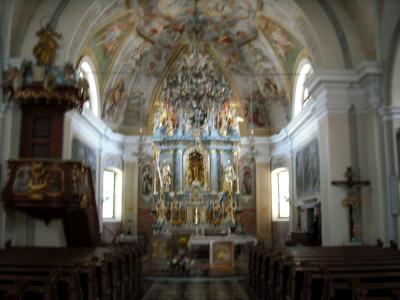 The interior of a Baroque church showing a highly ornate golden altar, a painted vaulted ceiling, a wooden pulpit on the left, and rows of pews.