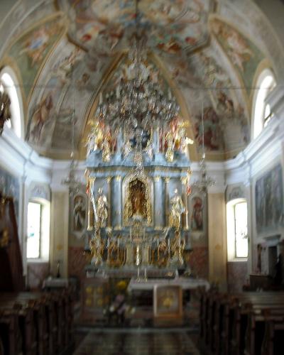 The interior of a Baroque church showing a highly ornate golden altar, a painted vaulted ceiling, a wooden pulpit on the left, and rows of pews.