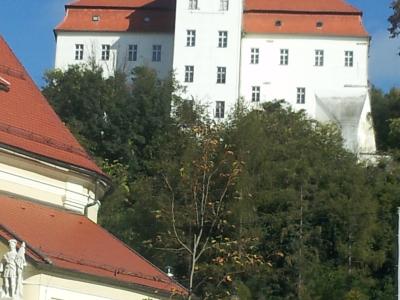 A large black wooden cannon carriage sits in the foreground on a grassy slope, with a long white building featuring a red-tiled roof, arched windows, and several flags (including the Slovenian flag) flying in front of it.