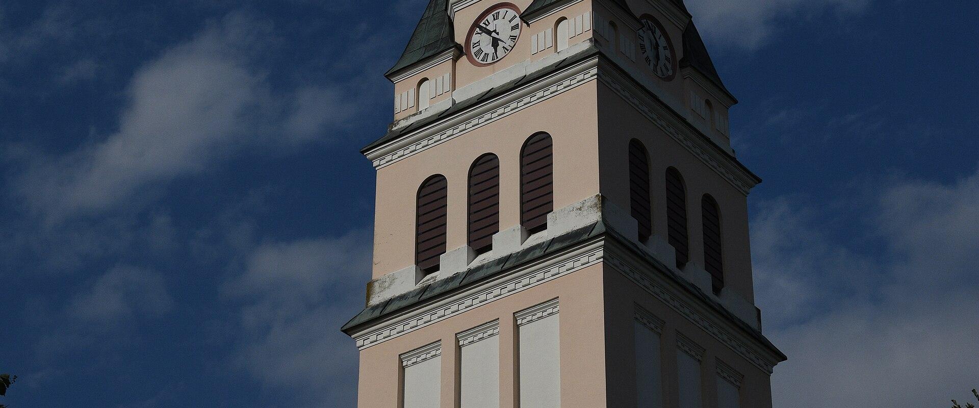 A low-angle shot of a tall church tower with a pale pink facade, white vertical pilasters, a dark green spire, and a clock face, set against a blue sky with clouds.