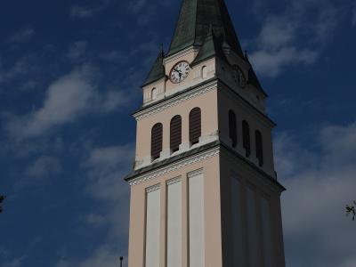 A low-angle shot of a tall church tower with a pale pink facade, white vertical pilasters, a dark green spire, and a clock face, set against a blue sky with clouds.