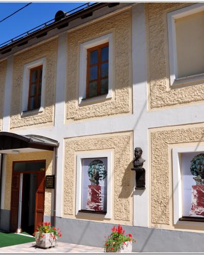 A two-story building with a textured yellow facade, white trim, and large display windows featuring posters for an exhibition titled "alojz gangl," with a small bust mounted on the wall and planters near the entrance.