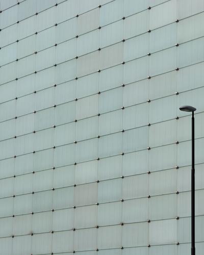 A close-up view of a modern building facade composed of a grid of light-colored rectangular panels with visible fasteners, and a single black street lamp post in the foreground.