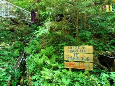 A wooden signpost in a lush forest setting with text reading "DEŽELA PRAVLJIC IN DOMIŠLJIJE" and "KOGA PRI CAROVNICI", alongside a figure dressed as a witch in the background.