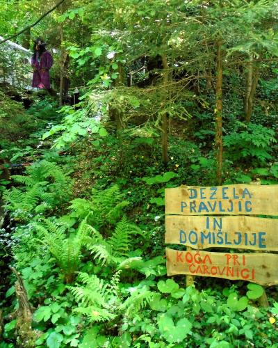 A wooden signpost in a lush forest setting with text reading "DEŽELA PRAVLJIC IN DOMIŠLJIJE" and "KOGA PRI CAROVNICI", alongside a figure dressed as a witch in the background.