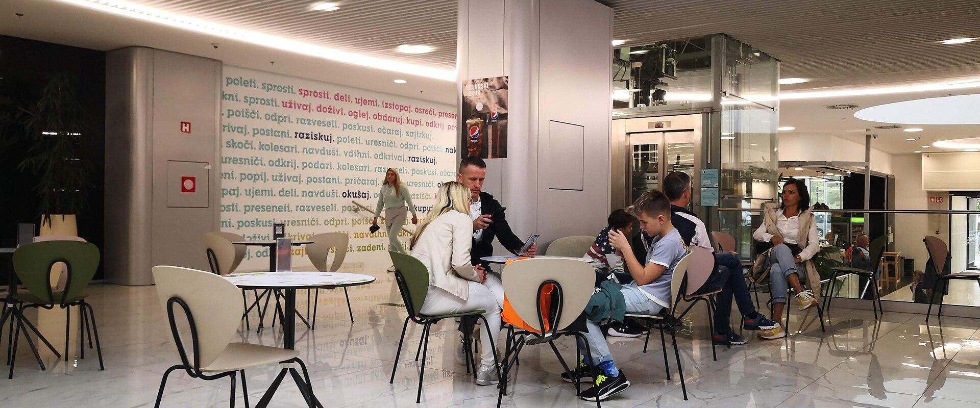 An indoor seating area with modern furniture, marble flooring, and a wall featuring Slovenian text, located within a commercial complex.