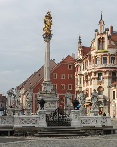 A tall stone plague column topped with a golden statue of the Virgin Mary, surrounded by a base with multiple stone statues, situated in a paved town square with historic buildings in the background.