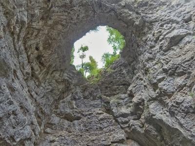 A low-angle view looking up through a rough, natural limestone archway towards a bright sky and green trees at the top of a rocky gorge.
