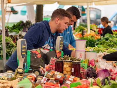 The image shows a close-up of a market stall with fresh produce, jars of preserved goods, and vendors interacting with customers under a tent.