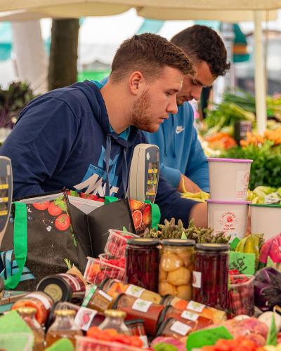 The image shows a close-up of a market stall with fresh produce, jars of preserved goods, and vendors interacting with customers under a tent.