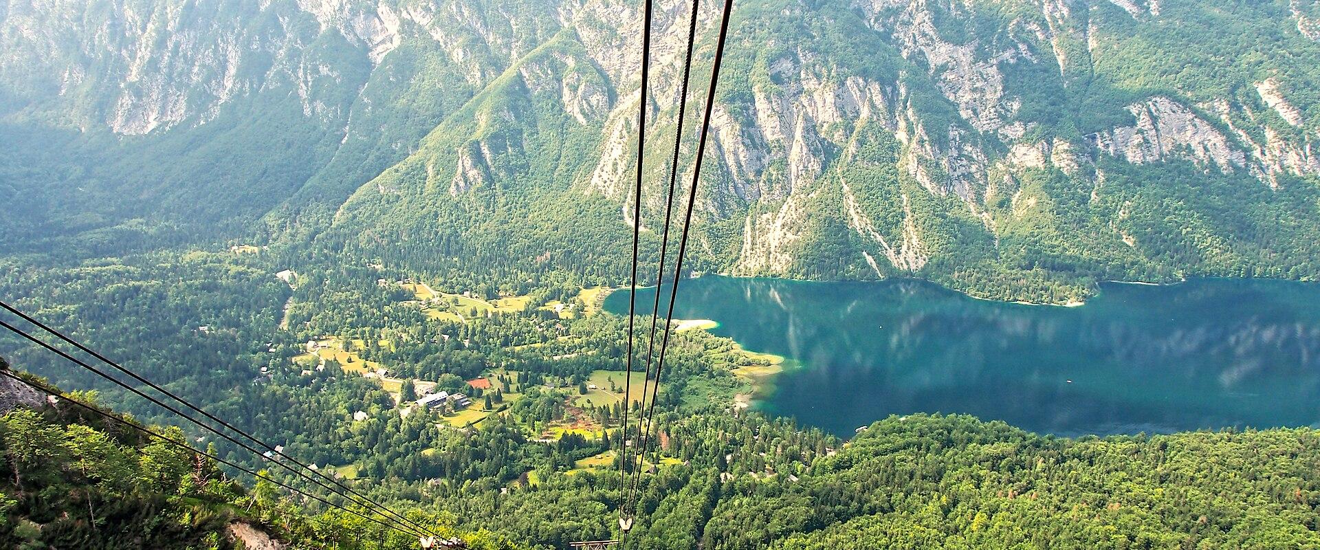 A high-angle panoramic photograph of a large, elongated alpine lake surrounded by steep, forested mountains and rocky cliffs under a partly cloudy sky.