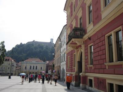 A wide pedestrian square with colorful historic buildings on the right, people walking, and Ljubljana Castle visible on a forested hill in the background.