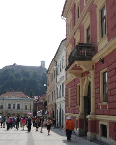A wide pedestrian square with colorful historic buildings on the right, people walking, and Ljubljana Castle visible on a forested hill in the background.