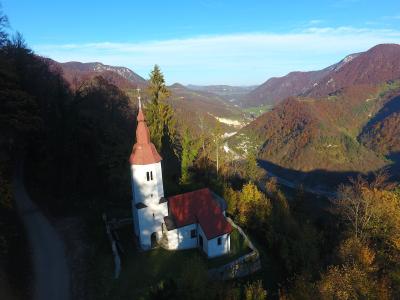 An aerial view of a white church with a distinctive red-tiled roof and a tall, bulbous steeple, situated on a grassy hillside overlooking a deep mountain valley with autumn foliage.