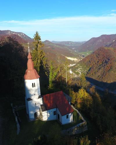 An aerial view of a white church with a distinctive red-tiled roof and a tall, bulbous steeple, situated on a grassy hillside overlooking a deep mountain valley with autumn foliage.