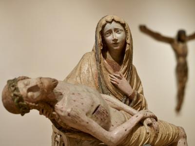 A polychrome wooden sculpture of the Pieta (Virgin Mary holding the dead Christ), with a blurred crucifix in the background, displayed indoors against a plain wall.