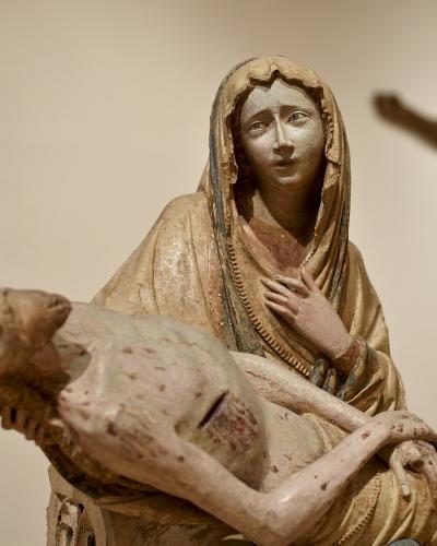 A polychrome wooden sculpture of the Pieta (Virgin Mary holding the dead Christ), with a blurred crucifix in the background, displayed indoors against a plain wall.