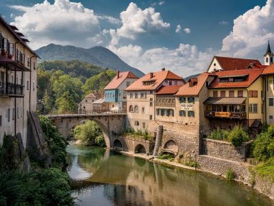 A stone arch bridge spanning a river in a historic European town, with colorful buildings lining the banks and mountains in the background; signage on a building reads 'HOTEL GARNI'.