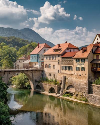 A stone arch bridge spanning a river in a historic European town, with colorful buildings lining the banks and mountains in the background; signage on a building reads 'HOTEL GARNI'.