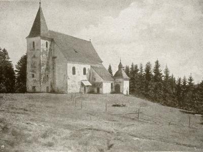 A black and white photograph of a stone church with a tall tower and a smaller adjacent chapel-like structure, situated on a grassy hill surrounded by a forest.
