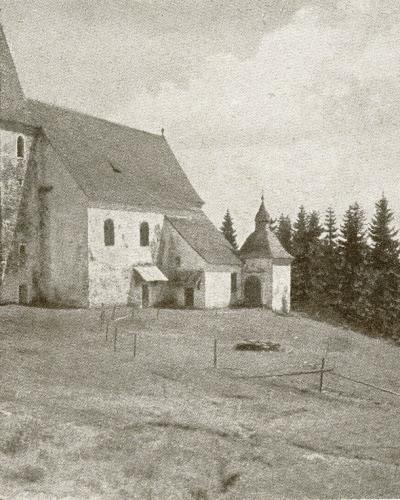 A black and white photograph of a stone church with a tall tower and a smaller adjacent chapel-like structure, situated on a grassy hill surrounded by a forest.