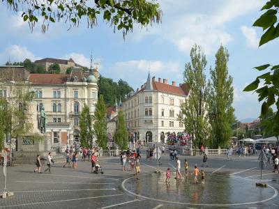 A wide-angle daytime photo of a large, paved public square with a central fountain where children are playing, surrounded by historic European-style buildings, a prominent statue on a pedestal to the left, and Ljubljana Castle visible on a hill in