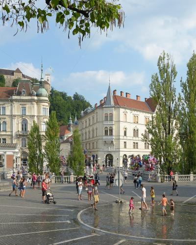 A wide-angle daytime photo of a large, paved public square with a central fountain where children are playing, surrounded by historic European-style buildings, a prominent statue on a pedestal to the left, and Ljubljana Castle visible on a hill in