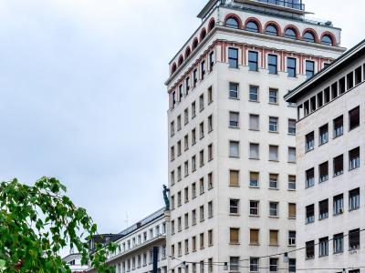 A low-angle photograph of the Nebotičnik skyscraper in Ljubljana, showing its Art Deco facade, distinctive corner windows, and rooftop structure against a cloudy sky, with trees and street-level elements in the foreground.