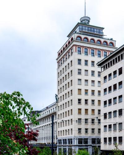 A low-angle photograph of the Nebotičnik skyscraper in Ljubljana, showing its Art Deco facade, distinctive corner windows, and rooftop structure against a cloudy sky, with trees and street-level elements in the foreground.