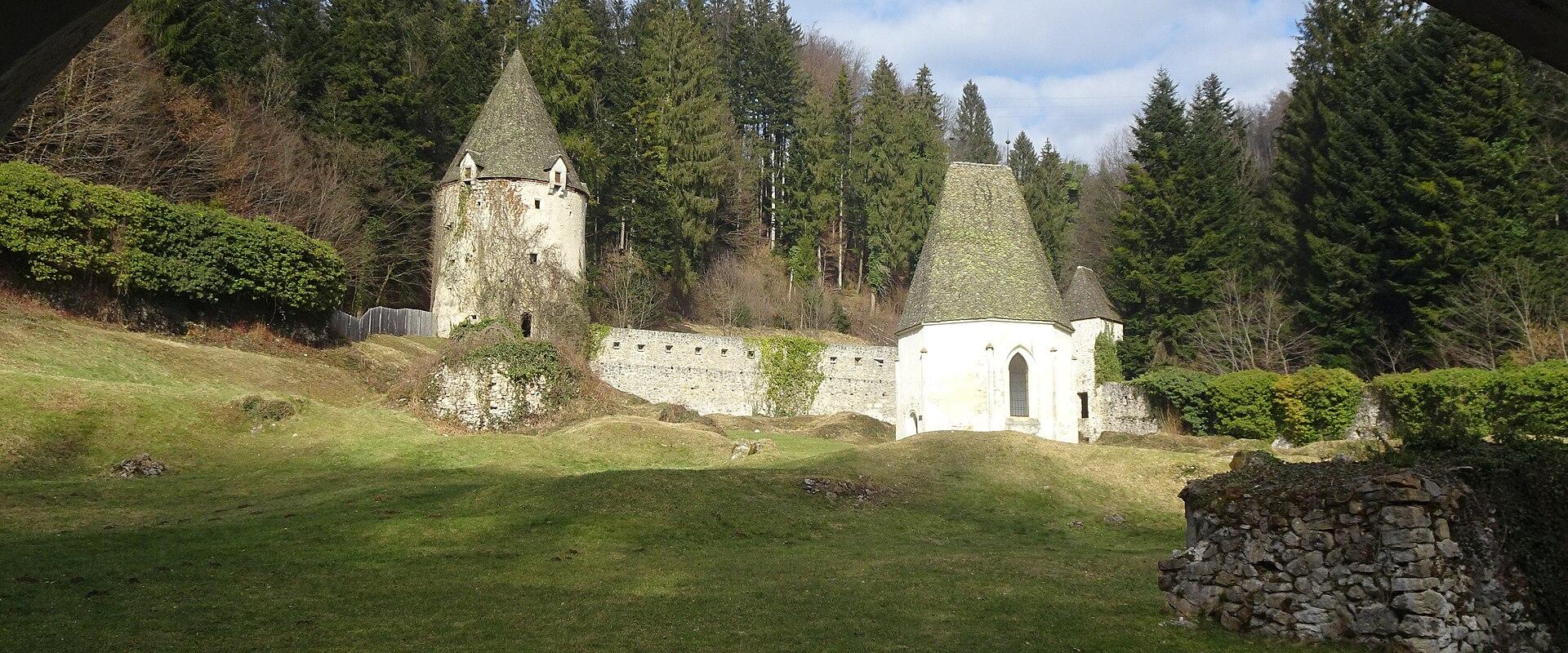 The image shows a view through a dark, arched opening of a white, cylindrical stone structure with a conical roof, situated on a grassy slope, with a forest in the background.