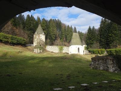 The image shows a view through a dark, arched opening of a white, cylindrical stone structure with a conical roof, situated on a grassy slope, with a forest in the background.