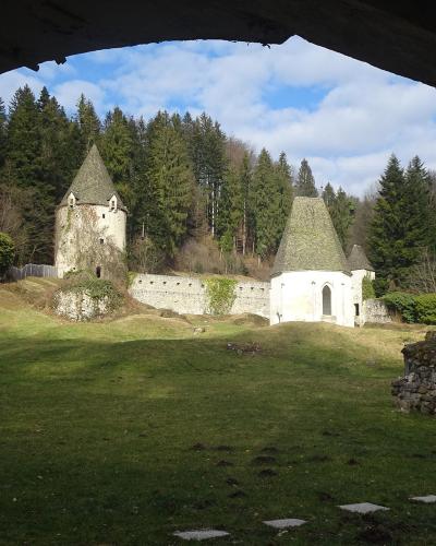 The image shows a view through a dark, arched opening of a white, cylindrical stone structure with a conical roof, situated on a grassy slope, with a forest in the background.