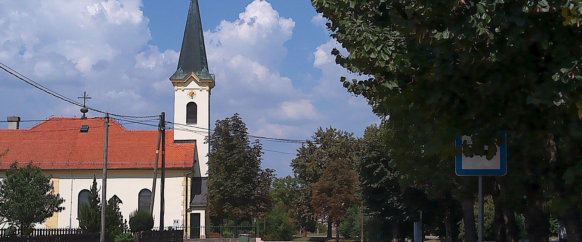 A white church with a red-tiled roof and a tall, dark spire stands beside a paved road under a blue sky with clouds, with trees and a bus stop sign visible in the foreground.