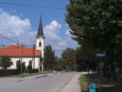 A white church with a red-tiled roof and a tall, dark spire stands beside a paved road under a blue sky with clouds, with trees and a bus stop sign visible in the foreground.