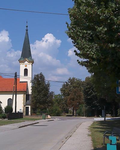 A white church with a red-tiled roof and a tall, dark spire stands beside a paved road under a blue sky with clouds, with trees and a bus stop sign visible in the foreground.