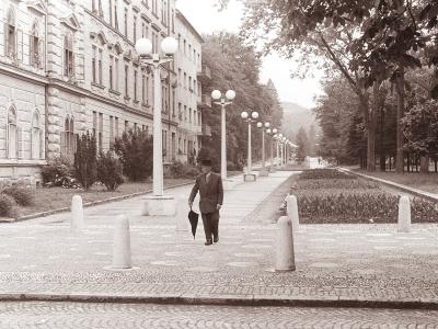 A black and white photograph showing a wide, paved promenade lined with distinctive spherical streetlights and bollards, with a large multi-story building on the left and a tree-lined park area on the right; a man in a suit and hat walks away from th