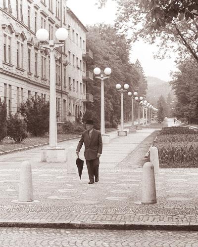 A black and white photograph showing a wide, paved promenade lined with distinctive spherical streetlights and bollards, with a large multi-story building on the left and a tree-lined park area on the right; a man in a suit and hat walks away from th
