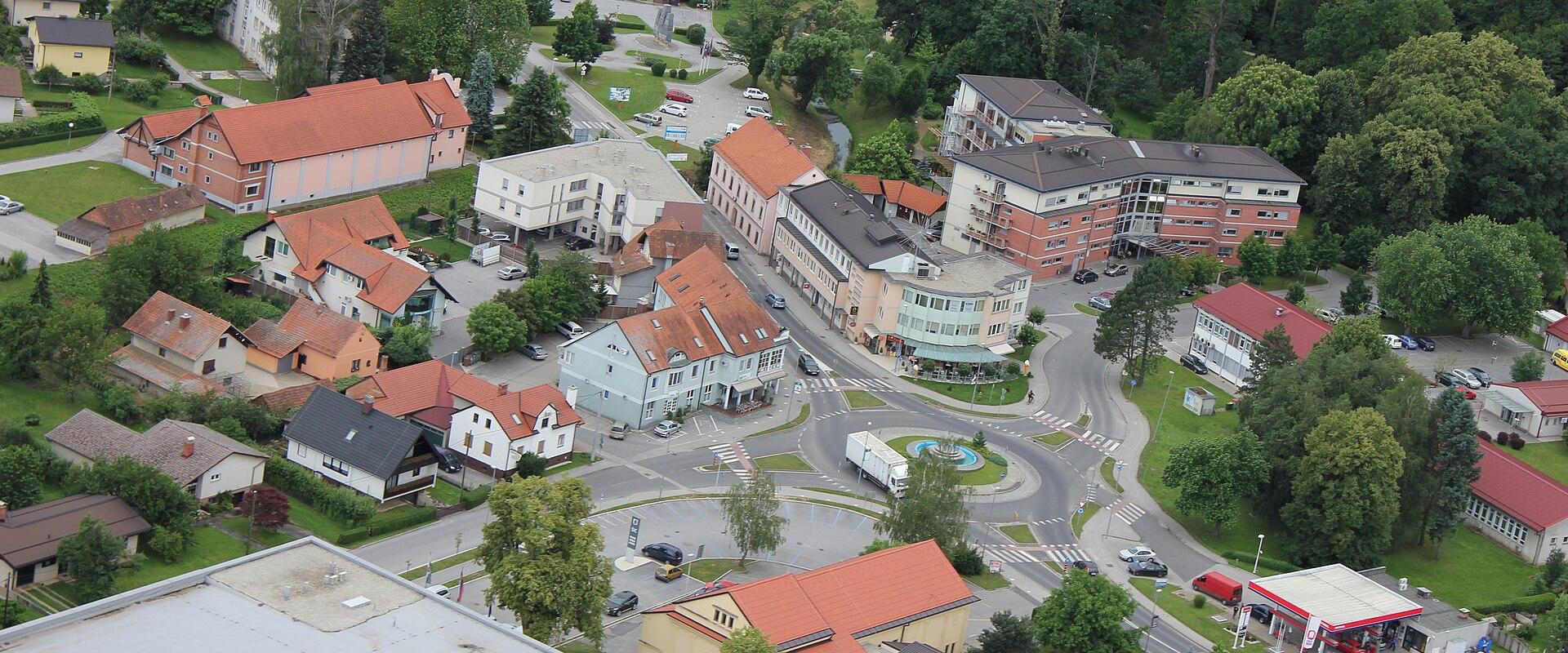 An aerial view of a town center featuring a roundabout, a gas station, and various residential and commercial buildings with red-tiled roofs.