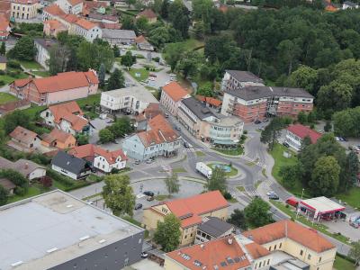 An aerial view of a town center featuring a roundabout, a gas station, and various residential and commercial buildings with red-tiled roofs.